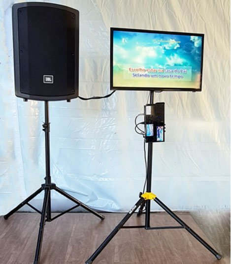 Karaokê montado com caixa JBL no tripé, TV e aparelho no suporte.